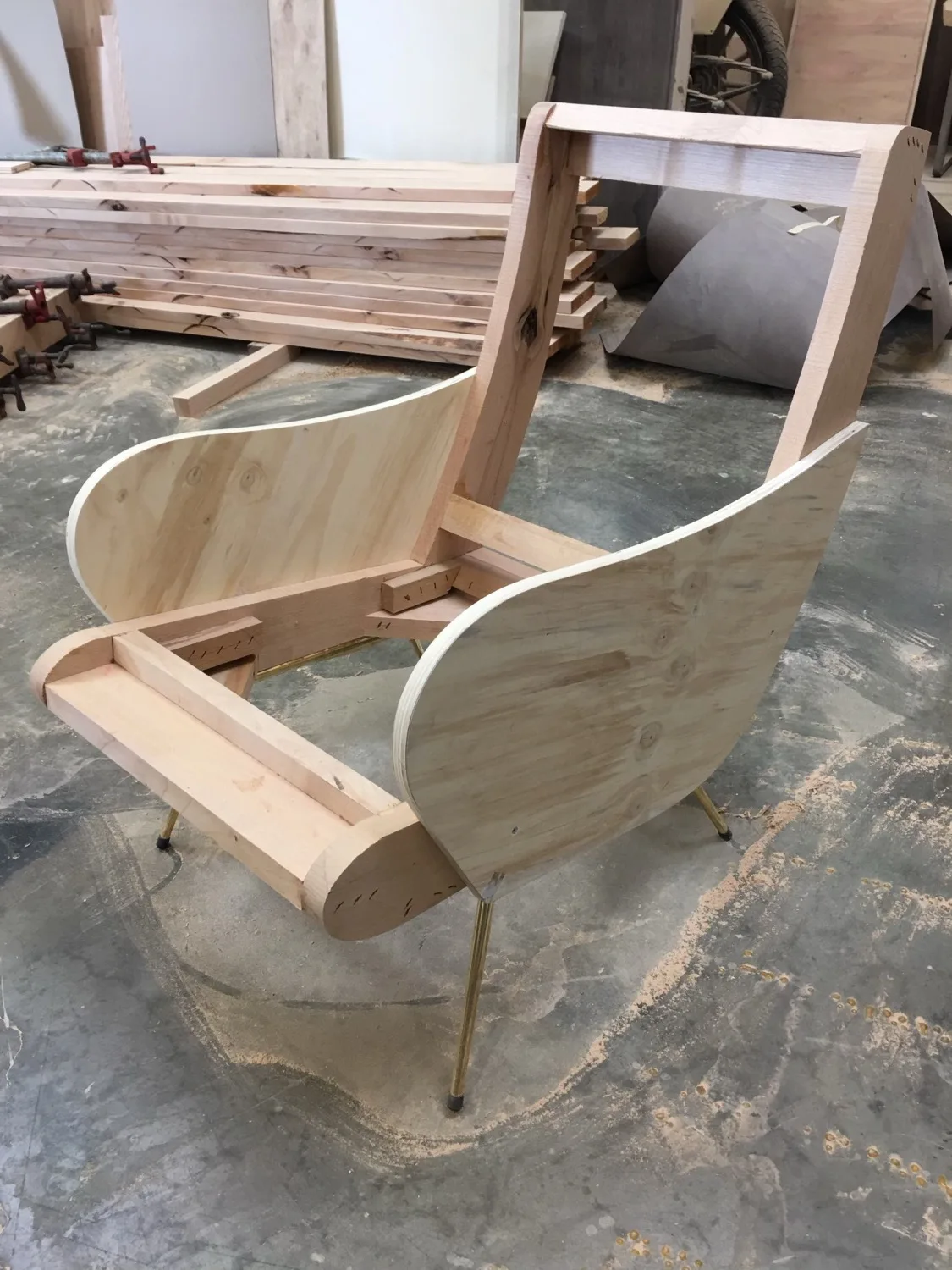 A curved hardwood lounge chair frame in progress on the shop floor, with brass legs set before upholstery